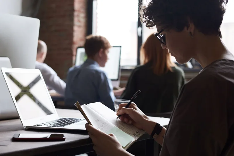 A person writing notes in a notebook at a desk with a laptop nearby, tracking job applications sent