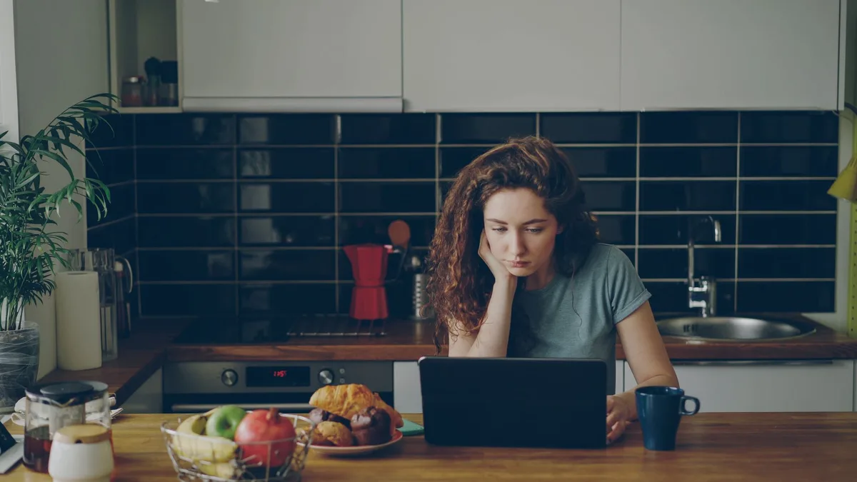 Woman thoughtfully reviewing something on her laptop at a home kitchen table