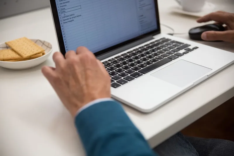 Laptop screen showing a spreadsheet with rows of data next to a small plate of crackers, representing the spreadsheet skills used in non-voice VA reporting work