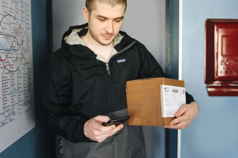 Man checking a phone while holding a labeled package, representing order tracking and fulfillment coordination