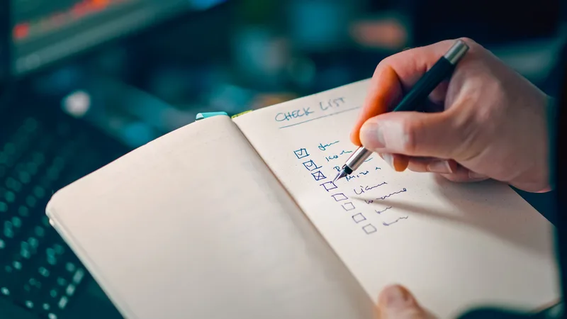 Close-up of a hand writing a checklist in a notebook beside a laptop keyboard