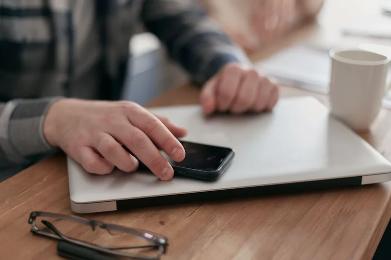 Hand resting on a smartphone placed on top of a closed laptop, with a mug and glasses nearby