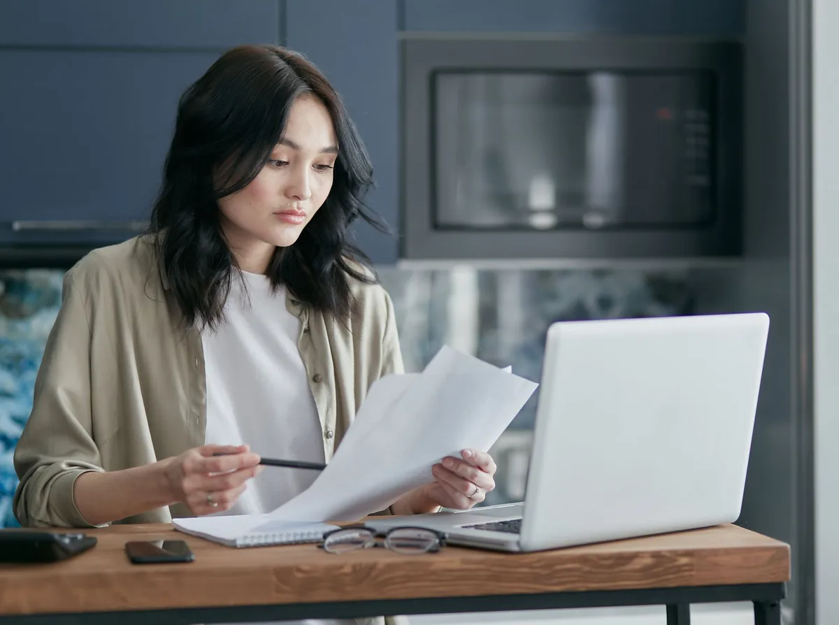 Woman reviewing papers next to an open laptop at a wooden table in a home kitchen
