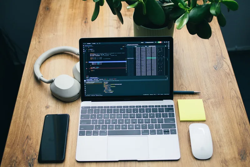 Laptop, headphones, smartphone, mouse, and sticky note arranged on a wooden desk with a plant